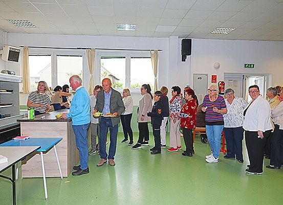 In einem Raum sind mehrere Menschen versammelt, einige stehen in einer Reihe. Ein Mann hält einen Teller mit Essen und ein anderer einen Teller. Es gibt ein Regal mit Gegenständen darauf und ein Tisch mit einem Buch darauf.