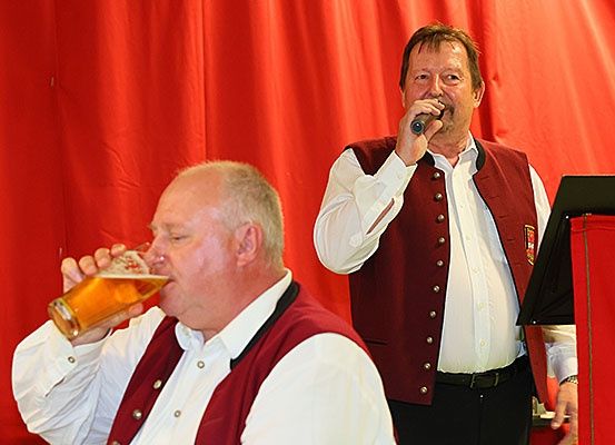 Zwei Männer auf der Bühne, einer trinkt ein Bier, während der andere in ein Mikrofon singt. Beide tragen traditionelle deutsche Kleidung.