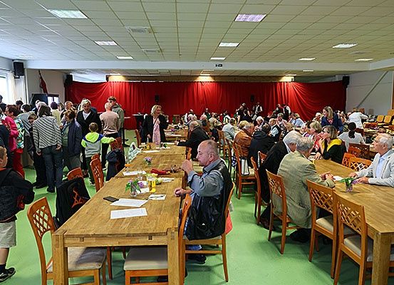 Eine große Gruppe von Menschen in einem Saal, um lange Tische herum sitzend, einige stehend, andere im Gespräch, mit roten Vorhängen im Hintergrund und einem grünen Boden.