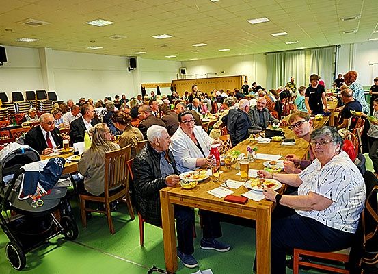 Eine große Gruppe von Menschen sitzt an langen Tischen in einem Speisesaal und isst Essen und Getränke. Einige Leute tragen Brillen und einige sind in Anzügen. Ein Kinderwagen steht im Vordergrund.