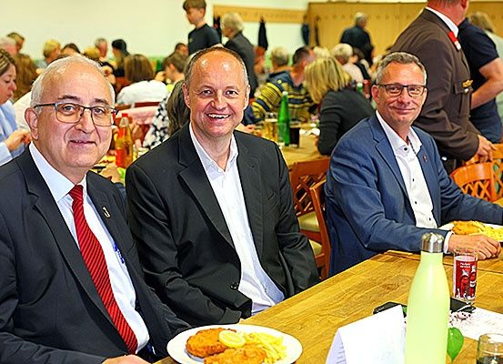 Drei Männer in Anzügen sitzen an einem Tisch in einem Restaurant. Sie lächeln und scheinen in ein Gespräch vertieft zu sein. Der Tisch ist mit Tellern, Gläsern und einer Flasche gedeckt.
