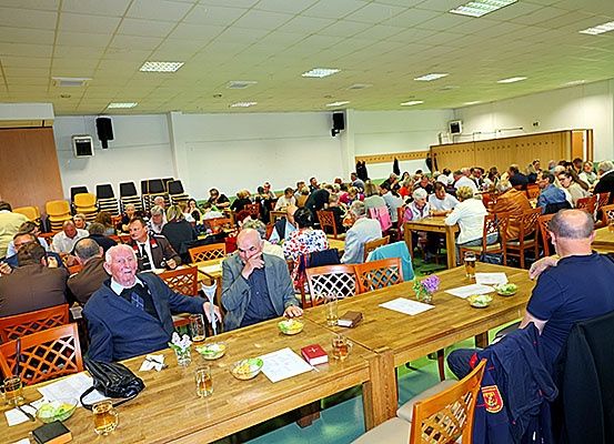 Ein großer Raum gefüllt mit Menschen, die an langen Holztischen sitzen, einige essen, andere unterhalten sich. Schüsseln, Gläser und Bücher sind auf den Tischen. Der Raum hat einen grünen Boden und weiße Wände.