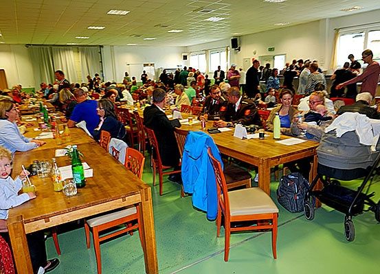 Ein großer Raum mit Tischen und Stühlen, in dem viele Menschen sitzen, einige mit Kindern. Flaschen, Gläser und andere Gegenstände stehen auf den Tischen. Ein Kinderwagen steht rechts, und eine Person steht in der Nähe einer Wand mit Fenster.