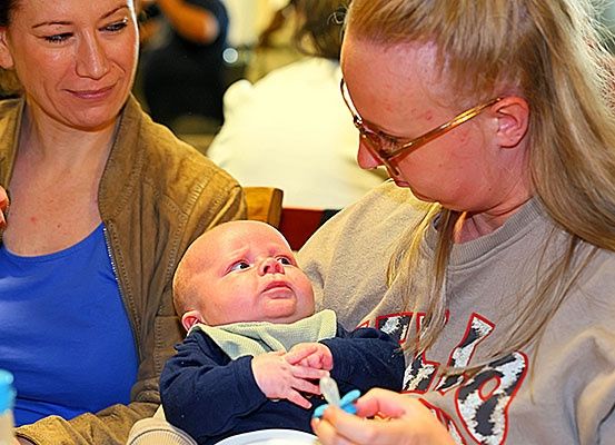 Zwei Frauen füttern ein Baby mit einem Löffel, eine hält das Baby, die andere einen Fläschchen.