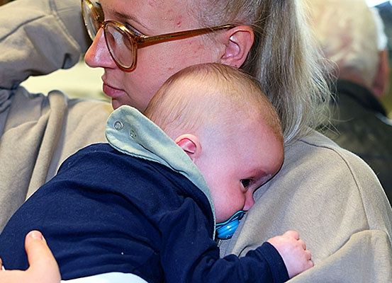 Eine Frau hält ein Baby in ihren Armen, mit einem Schnuller im Mund des Babys. Die Frau trägt eine Brille. Hinter ihnen sitzt ein älterer Mann.