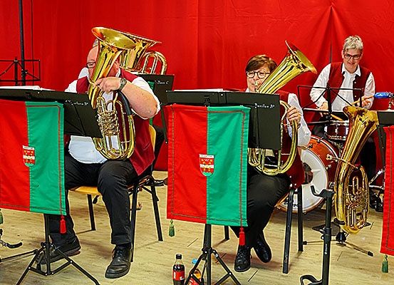 Drei Musiker sitzen auf Stühlen und spielen auf einer Bühne mit rotem Hintergrund Blechblasinstrumente. Sie tragen rote Westen.