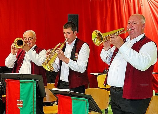 Drei Männer in roten Westen und weißen Hemden spielen Trompeten vor einem roten Hintergrund. Sie sind wahrscheinlich Teil eines Musikensembles.