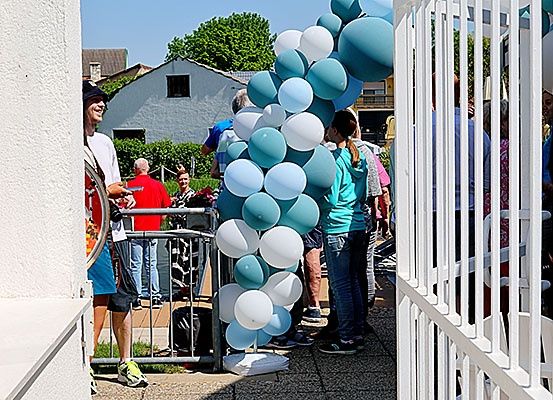 Eine Gruppe von Menschen steht draußen in der Nähe eines Gebäudes, einige warten in einer Schlange und andere schauen sich Luftballons an.