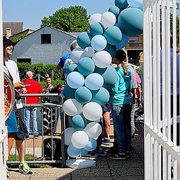 Eine Gruppe von Menschen steht draußen in der Nähe eines Gebäudes, einige warten in einer Schlange und andere schauen sich Luftballons an.