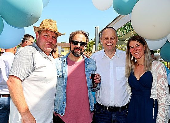 Vier Personen posieren für ein Foto vor blauen und weißen Luftballons. Ein Mann mit Hut, ein Mann in einer Jeansjacke, ein lächelnder Mann in einem weißen Hemd und eine Frau in einem blauen Kleid.