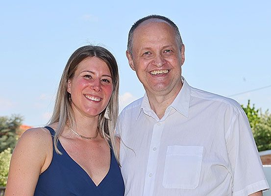 Ein Mann und eine Frau stehen eng beieinander, lächeln und posieren für ein Foto. Die Frau trägt ein blaues Kleid und der Mann ein weißes Poloshirt. Hinter ihnen sind einige Pflanzen und eine Mauer.