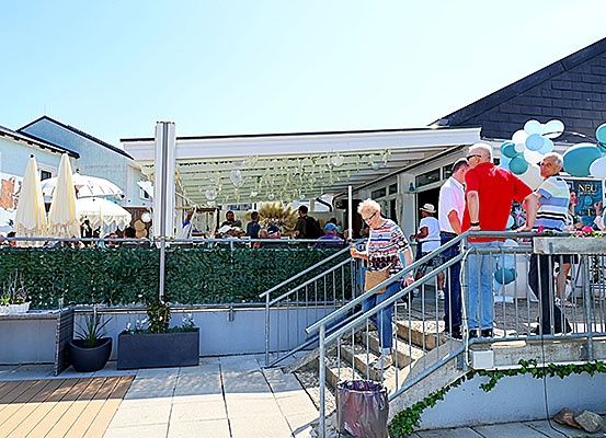 Ein Outdoor-Restaurant mit einem Vordach und Tischen mit sitzenden Menschen. Menschen stehen auf Treppen, eine mit einer Handtasche. Es gibt Topfpflanzen, Luftballons und Geländer.