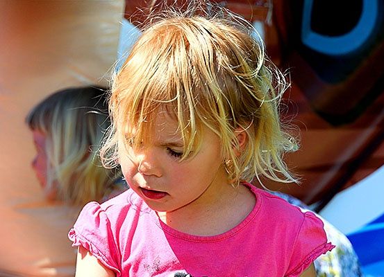 Ein junges Mädchen mit blonden Haaren, das ein rosa T-Shirt trägt, schaut nach unten in einem Außenbereich, möglicherweise einem Jahrmarkt, mit einem verschwommenen Bild eines anderen Mädchens im Hintergrund.