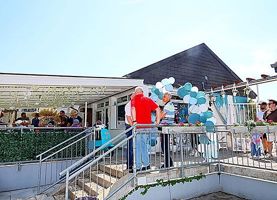 Menschen versammeln sich auf einer Terrasse vor einem Gebäude mit einer Treppe. Die Terrasse ist mit Geländern, Luftballons und Pflanzen dekoriert. Einige Leute tragen Hüte.