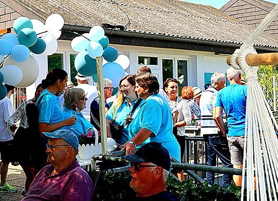 Eine Gruppe von Menschen in blauen T-Shirts versammelt sich im Freien, möglicherweise zu einer Feier, mit einem Haus mit Strohdach im Hintergrund. Viele Luftballons sind an einem Zaun befestigt.
