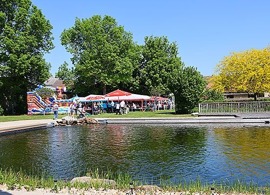 Ein Park mit einem Teich, umgeben von Bäumen und Büschen, mit Menschen und einem Zelt, das sich am Wasser versammelt.