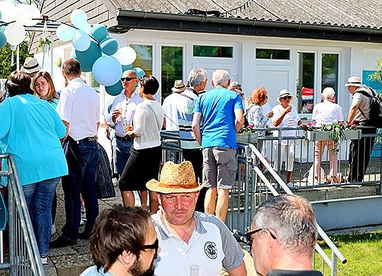 Eine Gruppe von Menschen steht vor einem weißen Gebäude mit Ballons, einige tragen Hüte und Brillen. Das Gebäude hat Glasfenster und Türen.