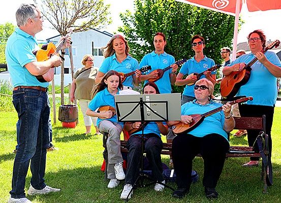 Eine Gruppe von Menschen spielt verschiedene Musikinstrumente an einem sonnigen Tag. Einige stehen, andere sitzen auf einer Bank. Sie tragen alle blaue Hemden.