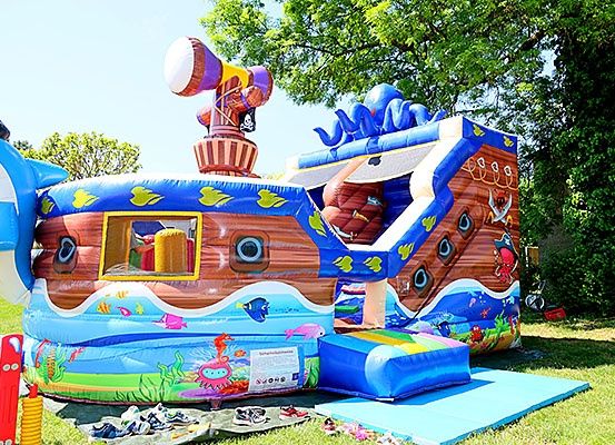 Ein großer aufblasbarer Piratenschiff mit Rutsche und Kanone steht in einem Park. Bäume und Gras umgeben das Gebiet.