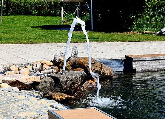 Ein Wasserbrunnen spritzt Wasser in einen von Steinen und Grünpflanzen umgebenen Teich. Der Brunnen befindet sich auf einem gepflasterten Bereich.
