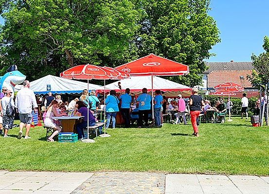 Outdoor-Veranstaltung mit Menschen unter Zelten. Einige sitzen, andere gehen. Rote Schirme und ein großer aufblasbarer Hai im Hintergrund.