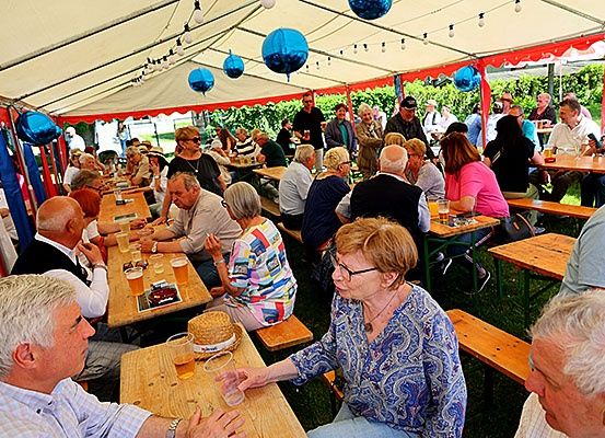 Eine Gruppe von Menschen sitzt an Tischen in einem Zelt, einige mit Brille, und trinken aus Bechern. Die Tische sind mit Essen und Getränken gefüllt. Blaue Luftballons hängen von der Decke.