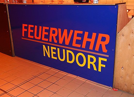 Ein blaues Schild mit orange und gelber Schrift. Das Schild trägt in fetten Buchstaben 'Feuerwehr Neudorf'.