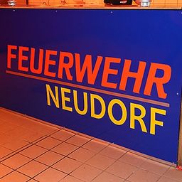 Ein blaues Schild mit orange und gelber Schrift. Das Schild trägt in fetten Buchstaben 'Feuerwehr Neudorf'.