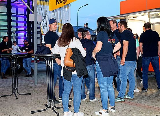 Mehrere Menschen stehen draußen, möglicherweise in einer Schlange vor einem Imbisswagen. Eine Frau in einem weißen Shirt ist unter ihnen.