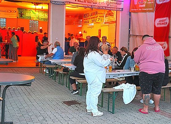 Eine Gruppe von Menschen isst in einem Außenrestaurant mit Tischen und Bänken. Der Bereich ist mit warmer Beleuchtung beleuchtet. Es gibt Banner und Schilder an der Wand.