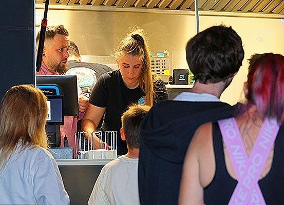 Eine Frau in einem schwarzen T-Shirt arbeitet an einem Essensausgabeschalter. Einige Leute warten in der Schlange, darunter ein Mann mit einem Tattoo auf dem Rücken.