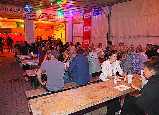 Menschen essen bei Nacht draußen unter einem Baldachin. Verschiedene Getränke und Speisen stehen auf den Tischen. Es gibt Werbung für Ford, Suzuki und Weingrill.