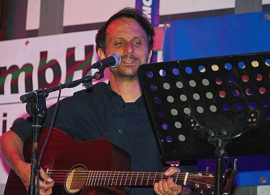 Ein Mann spielt Gitarre und singt vor einem Mikrofon auf der Bühne, mit einem Notenständer dahinter.