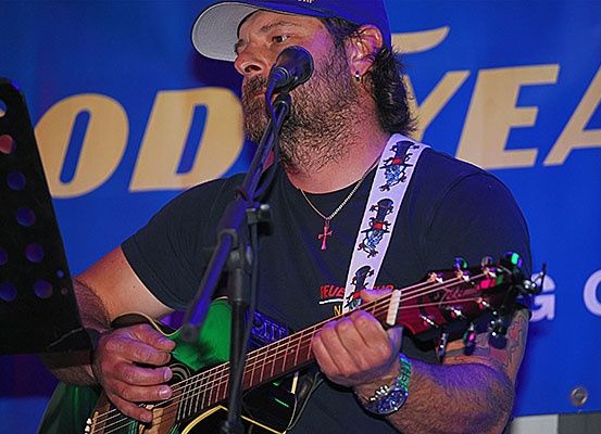 Ein bärtiger Mann spielt Gitarre vor einem Mikrofon, wahrscheinlich bei einem Konzert.