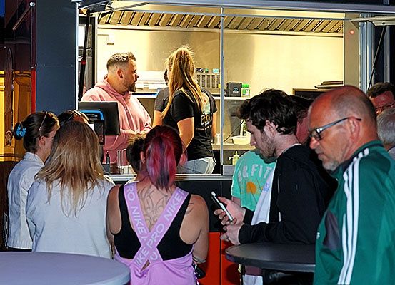 Eine Gruppe von Menschen steht bei Nacht vor einem Imbisswagen. Im Inneren spricht ein Mann in einem rosa Shirt mit einer Frau mit langen Haaren. Eine andere Frau trägt eine Schärpe, auf der 'Nike Pro' steht.