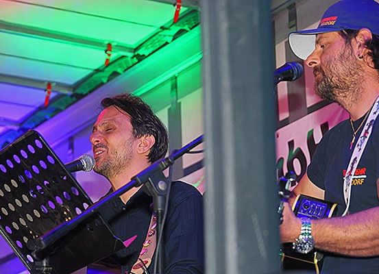 Zwei Männer treten auf der Bühne auf, einer singt in ein Mikrofon und der andere spielt Gitarre. Bunte Lichter beleuchten den Hintergrund.