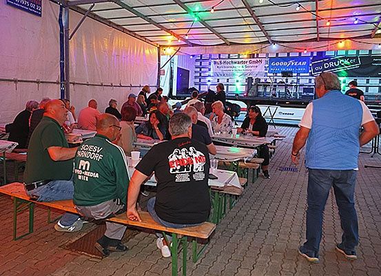Eine Gruppe von Menschen sitzt an Tischen bei einer Outdoor-Veranstaltung, möglicherweise einem Musikfestival. Ein Mann geht an ihnen vorbei, und eine Bühne ist hinter ihnen.