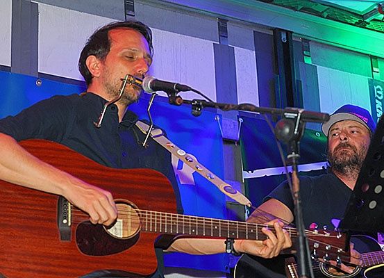 Zwei Männer spielen auf der Bühne mit Gitarren, ein Mann spielt Mundharmonika in ein Mikrofon.