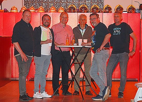 Fünf Männer stehen hinter einem kleinen Tisch in einem hell beleuchteten Raum, in lässiger Kleidung. Einer hält einen Becher, und zwei haben Flaschen vor sich. Hinter ihnen sind rote Wände und Helme zu sehen.