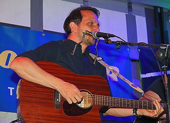 Ein Mann spielt Gitarre und singt in ein Mikrofon. Er hat einen Gurt um den Hals. Hinter ihm steht eine Person, die Gitarre spielt. Es gibt ein blaues Banner mit dem Wort 'TE'.