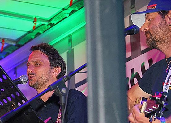 Zwei Männer spielen auf der Bühne mit Gitarren, einer singt in ein Mikrofon und der andere streicht seine Gitarre. Sie werden von grünen Lichtern beleuchtet.