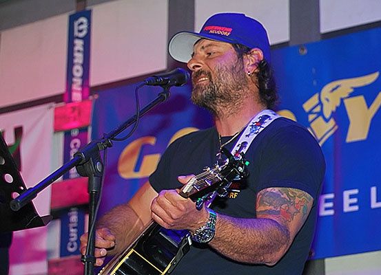 Ein Mann mit Tattoos am Arm singt in ein Mikrofon und spielt Gitarre.