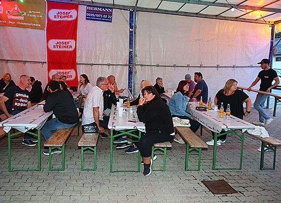 Eine Gruppe von Menschen sitzt um Tische unter einem Zelt, einige mit Getränken und Essen, während ein Mann in der Nähe eines Tisches steht und ein Auto draußen geparkt ist.