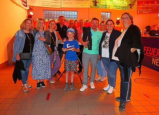 Eine Gruppe von Menschen, einschließlich Erwachsener und Kindern, steht lächelnd in einem Saal, wobei ein Junge eine Pizza hält.
