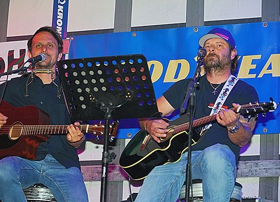 Zwei Männer spielen Gitarre und singen auf der Bühne, einer benutzt eine Mundharmonika. Hinter ihnen ist ein blauer Banner mit gelbem Text.