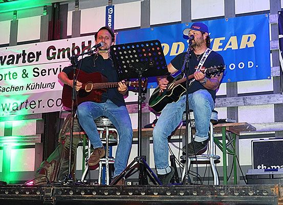 Zwei Männer spielen auf der Bühne mit Gitarren, einem Mikrofon und einem Notenständer. Sie tragen Freizeitkleidung und haben ein Banner mit Logo und Text hinter sich.