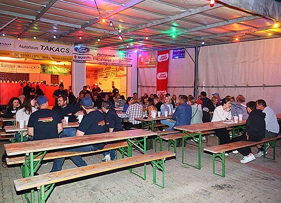 Ein überfüllter Außenbereich mit Tischen und Bänken, Menschen sitzen und stehen, manche tragen Hüte. An der Decke hängen Werbetafeln, darunter Renault, Ford und Suzuki.