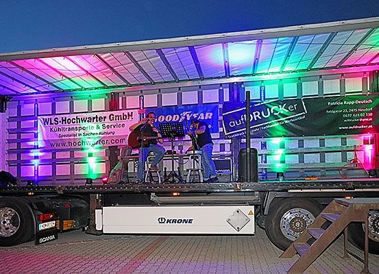 Zwei Männer treten auf einer Bühne auf, die auf einem Anhänger aufgebaut ist, mit bunten Lichtern, einem Hintergrundbanner mit der Aufschrift 'WLS-Hochwarter GmbH' und einem Mikrofonständer. Die Bühne befindet sich auf einem Anhänger mit einem 'Krone'-Logo an der Seite.