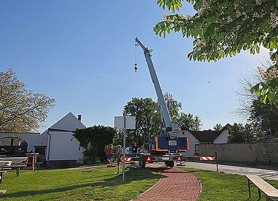Ein Kran steht in einem grasbewachsenen Bereich mit einem Weg, umgeben von einem weißen Gebäude, Bäumen und einer Bank. Arbeiter bedienen den Kran.