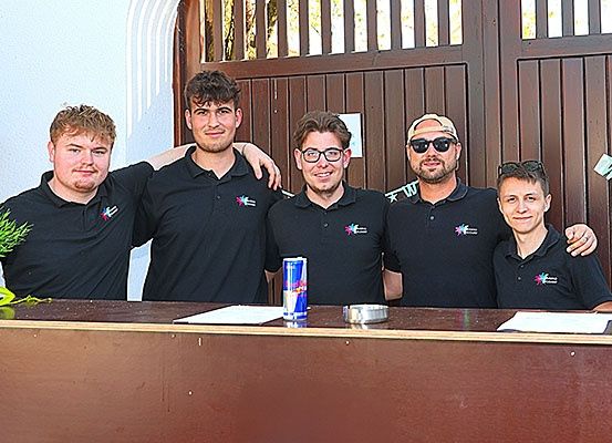 Eine Gruppe von Männern in schwarzen Poloshirts lächelt und posiert für ein Foto an einer Bartheke mit einer Pflanze, einer Dose Red Bull und einigen Papieren.